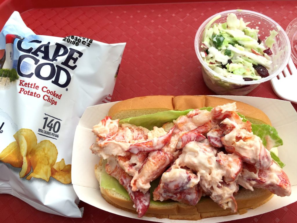 Small bag of Cape Cod Potato Chips, lobster roll in a bun in a holder, and side of coleslaw on a red tablecloth