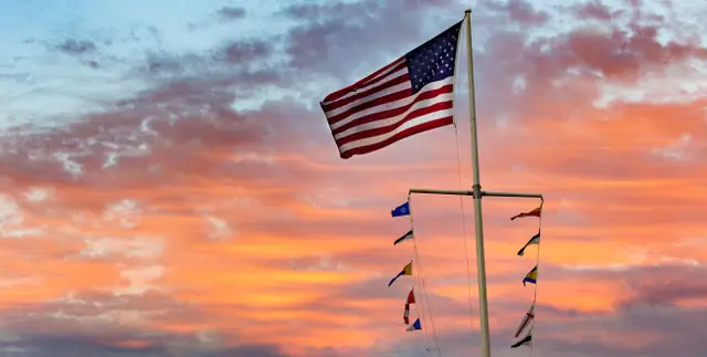 American flag flying on a tall flagpole against a colorful sunset sky with shades of orange, pink, and blue, creating a patriotic and scenic coastal atmosphere.