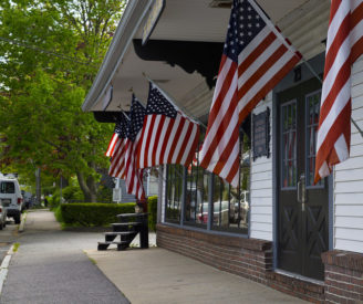 American flags hanging off the exterior of a white building with an overhang next to the sidewalk | Brewster By the Sea Cape Cod Inn | Brewster, MA