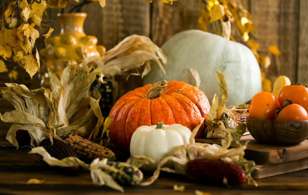 A festive Thanksgiving or fall harvest display featuring an orange pumpkin, a small white pumpkin, decorative corn cobs, dried corn husks, a light blue-green squash, and a bowl of persimmons, all set on a rustic wooden table.