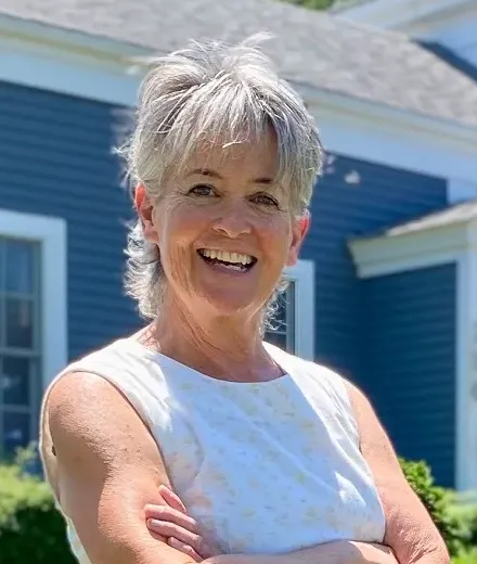 Smiling woman with short gray hair standing outdoors with arms crossed, wearing a sleeveless white top in front of a blue house.