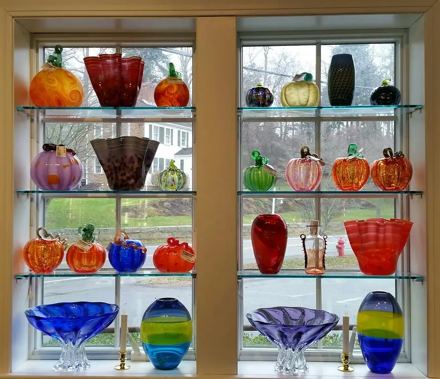 A display of colorful glass art pieces arranged on shelves, featuring various shapes such as bowls, vases, and decorative pumpkins, against a window backdrop.