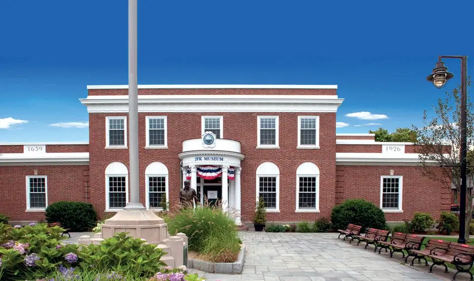 A brick building with white accents and flags, featuring a round entrance, surrounded by greenery and benches.